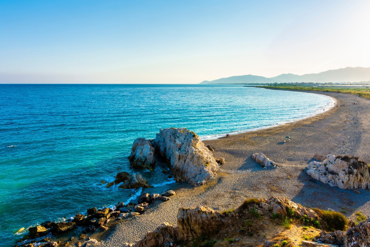 Eine weite Küstenlandschaft mit türkisblauem Meer und einem geschwungenen Kiesstrand. Im Vordergrund sind kleine Felsen, im Hintergrund grüne Felder und Berge zu sehen.