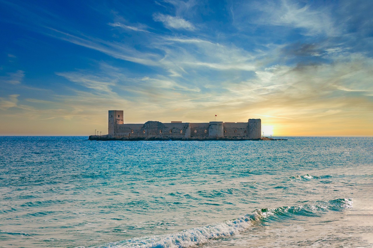Sonnenuntergang über dem Meer mit einer mittelalterlichen Burg auf einer kleinen Insel vor der Küste.
