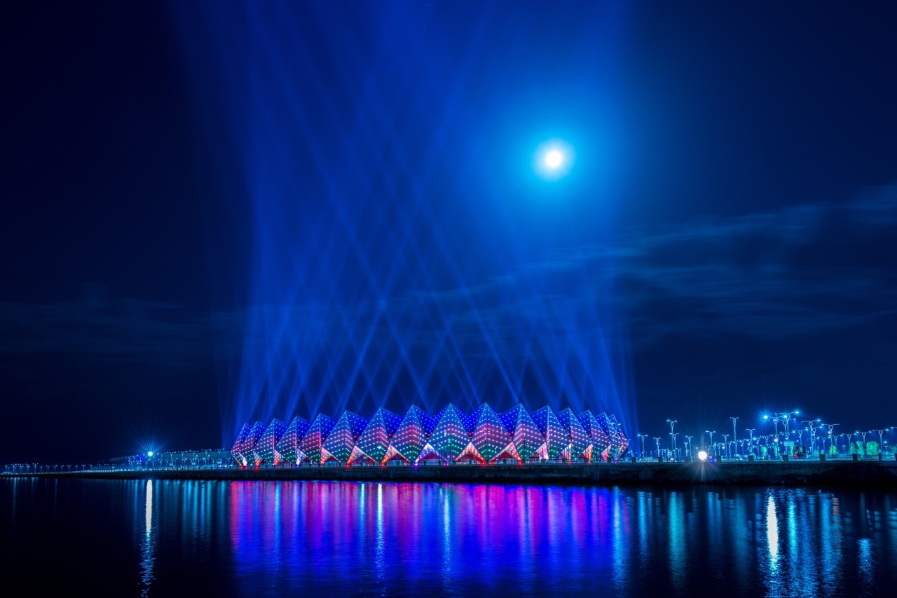 Im Zentrum des Bildes befindet sich ein modernes, kreisförmiges Gebäude bei Nacht, das in blau und pink leuchtet. Die Lichtstrahlen erheben sich zum Himmel und spiegeln sich im Wasser vor dem Gebäude. Der Bau setzt sich aus einzelnen kegelförmigen Bauteilen zusammen.