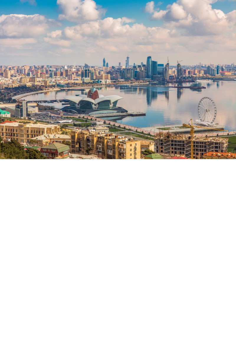 Luftansicht einer Küstenstadt, die sich im Halbkreis um eine Meeresbucht erhebt. Im Hintergrund ist eine Skyline mit Wolkenkratzern zu sehen, im Bildzentrum ein moderner Bau mit einer blütenförmigen Dachkonstruktion, der ins Wasser hineinragt. Im Vordergrund sind mehrstöckige Häuser und ein Riesenrad sichtbar.