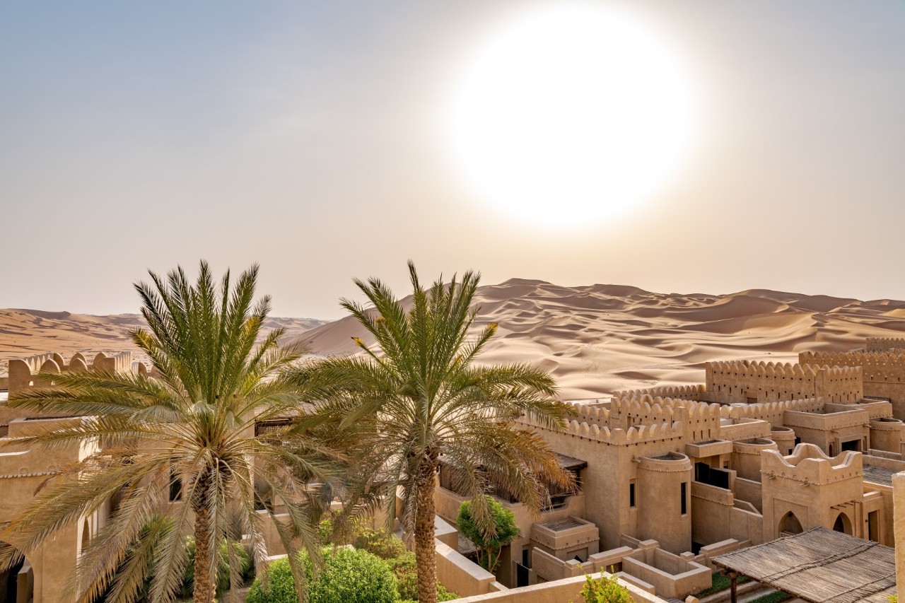 Das Bild zeigt traditionelle arabische sandfarbene Häuser mit Mauern, eingebettet in eine Oase mit Palmen. Im Hintergrund erstrecken sich weite Sanddünen unter der strahlenden Sonne.