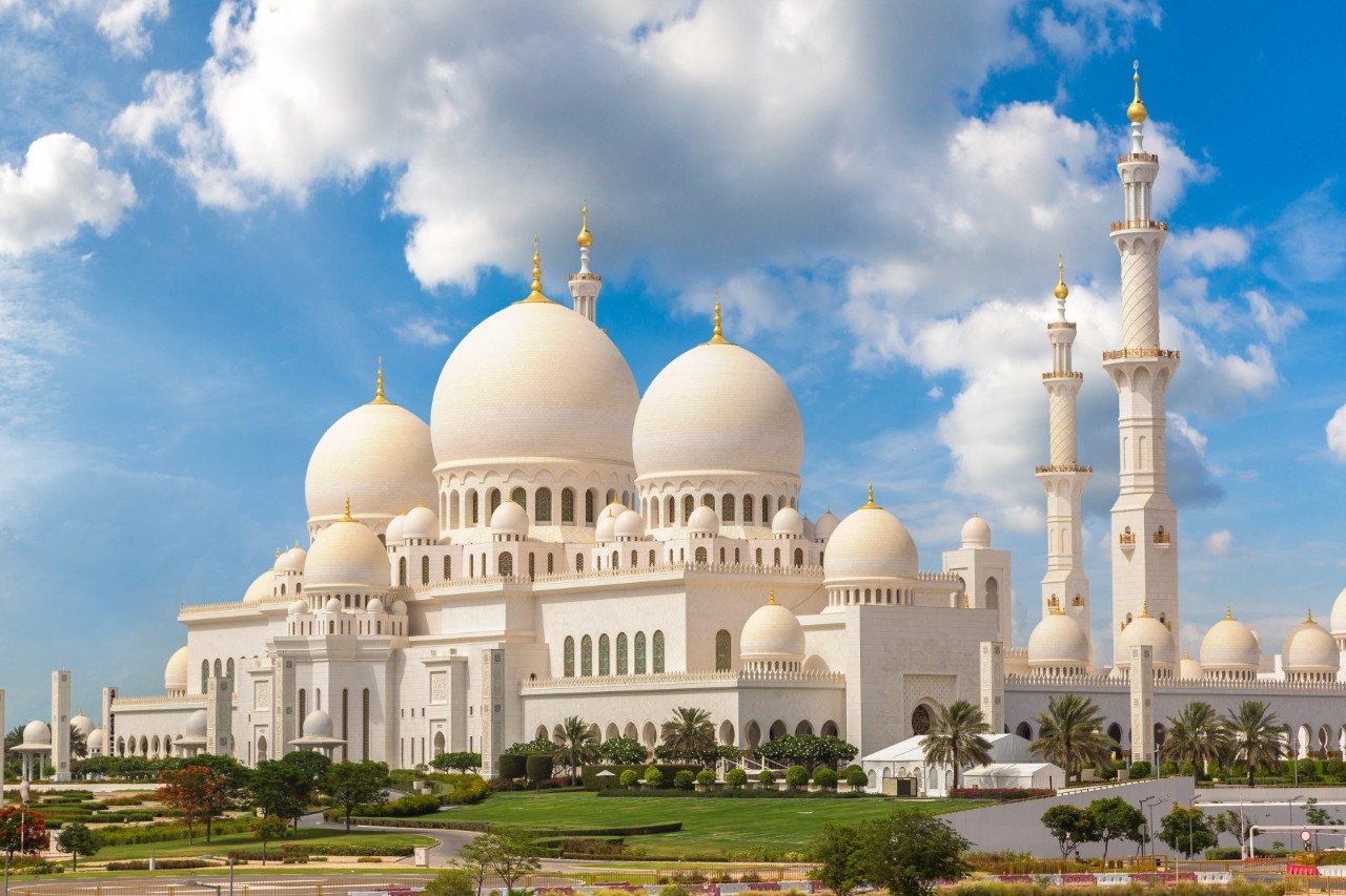 Das Bild zeigt eine beeindruckende Moschee mit weißen Marmorkuppeln, kunstvollen Minaretten und detaillierter islamischer Architektur, die von einer grünen Gartenlandschaft umgeben ist. 