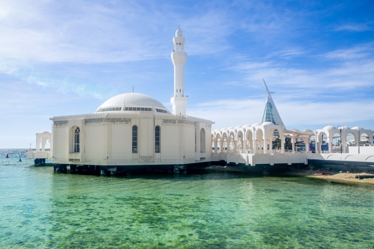 Schwimmende, weiße Moschee mit Minarett auf türkisblauem Wasser. 