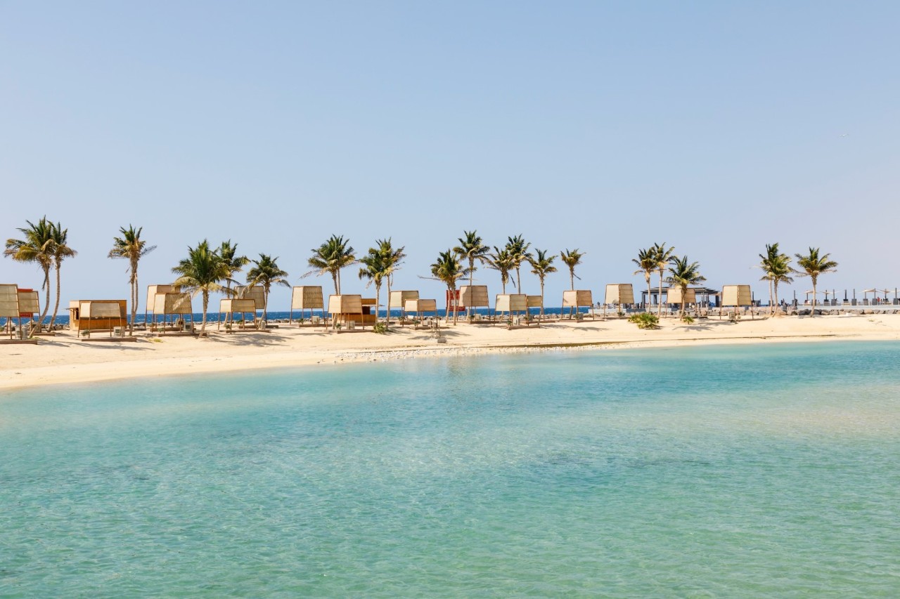 Klares, blaues Wasser auf einen weißen Sandstrand mit Palmen und Strandkabinen.  