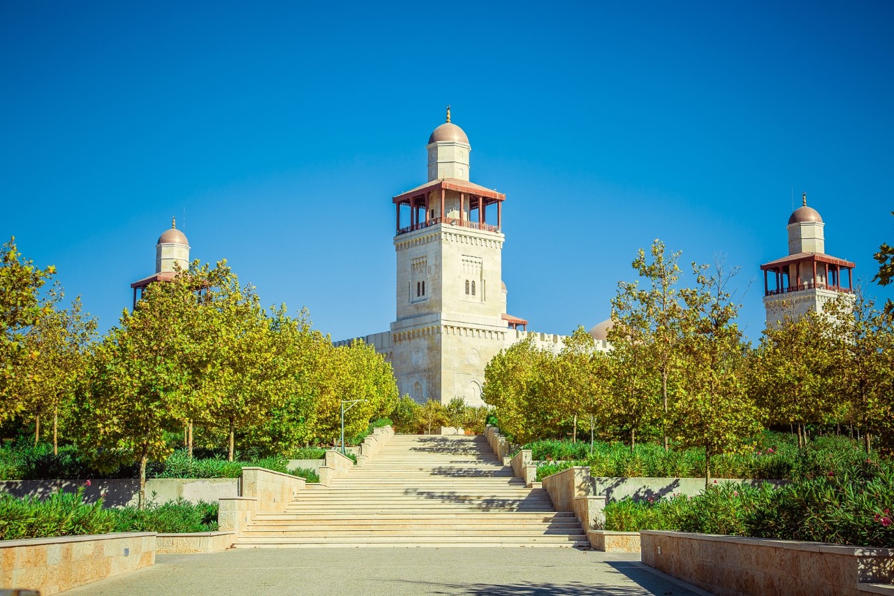 Blick auf eine weiße Mauer mit vier Türmen in jeder Ecke, die als Minarette dienen und auf denen je sich jeweils eine Kuppel befindet. Dahinter ist nicht sichtbar im Bild die Moschee. Zur Anlage führt eine breite Treppe, neben der sich links und rechts Blumen und Bäume befinden.