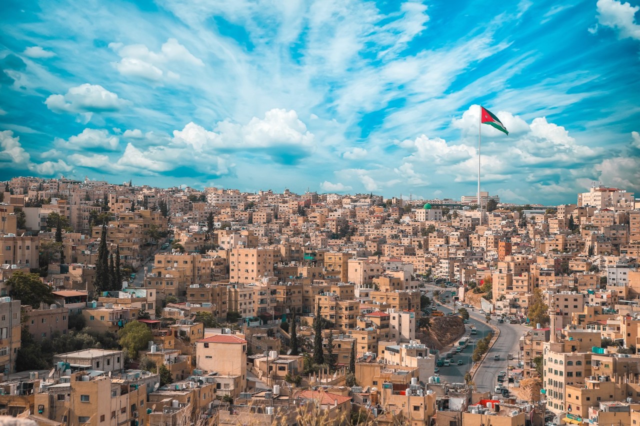 Blick von oben auf eine dicht bebaute Stadt mit beige bis sandfarbenen Häusern und einer Straße, die sich rechts durch die Stadt schlängelt. Rechts im Bild ragt eine riesige jordanische Fahne in den bewölkten Himmel.