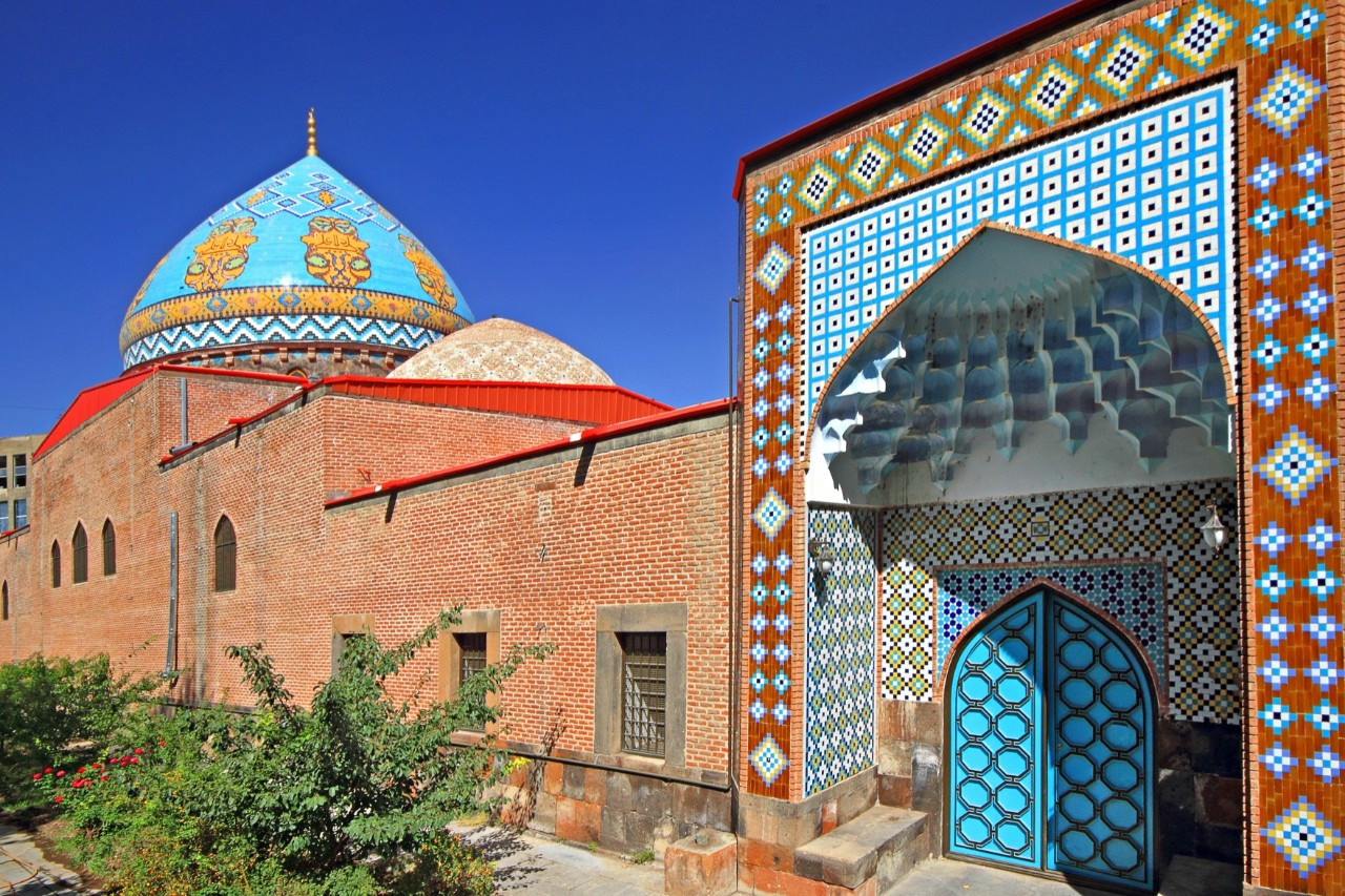 Seitlicher Blick auf eine Moschee mit orange-blau verziertem Kuppeldach und mosaikartigen Wandverzierungen im Eingangsbereich auf der rechten Bildseite. Das Hauptgebäude besteht aus rötlichen Backsteinen.