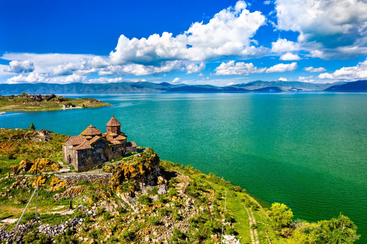 Blick von oben auf einen großen, grünblauen See, an dessen linker Uferseite ein Kloster zu sehen ist. Im Hintergrund zeichnet sich ein flaches Gebirgspanorama ab. Über dem See sind Wolken am blauen Himmel zu sehen.