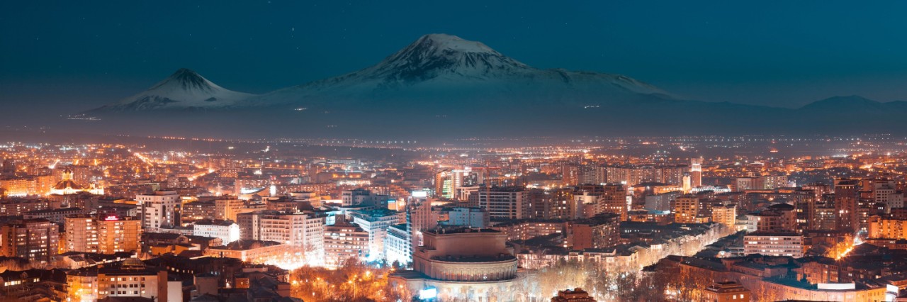 Blick von oben auf eine hell beleuchtete Stadt in der Nacht mit schneebedeckten Bergen im Hintergrund und dunkelblauem Nachthimmel.