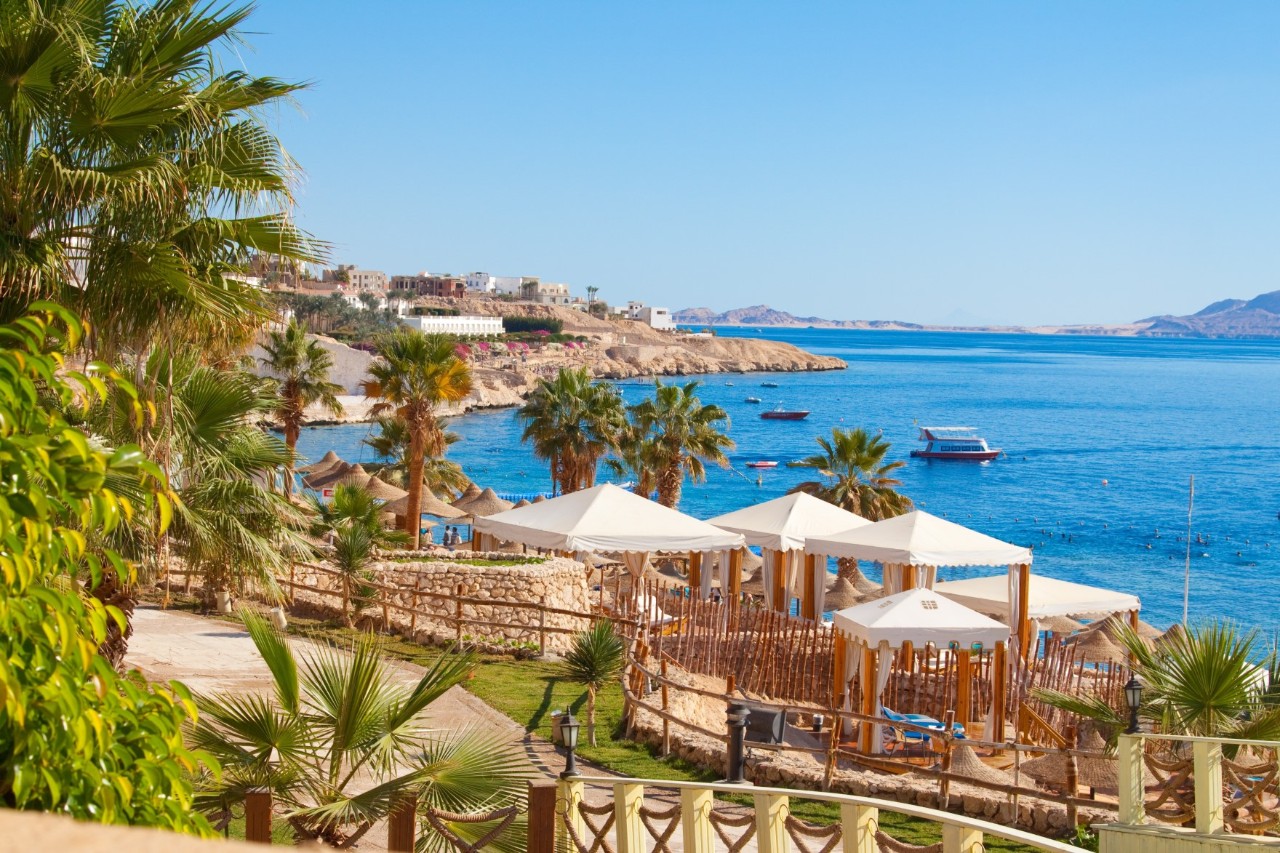 Ferienresort mit Palmen und Sonnenbetten an der Küste, Meer, Boote und Felsen im Hintergrund  