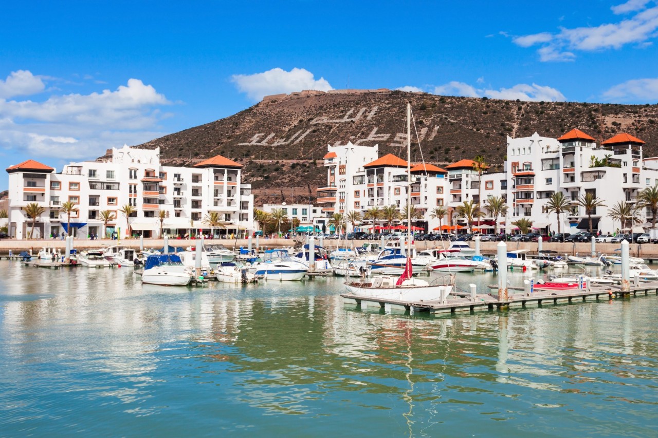 Blick auf die Marina von Agadir, einen kleinen Hafen, in mit einer Vielzahl von geparkten Booten. Die Marina ist umgeben von modernen weißen Gebäuden mit roten Dächern. Im Hintergrund erhebt sich ein Hügel mit einem in den Felsen gehauenen Schriftzug in arabischer Schrift, der die Worte "Gott, König, Vaterland" darstellt. Der Himmel ist klar und blau, was die sonnige Atmosphäre unterstreicht.