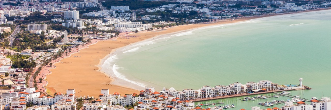 Luftaufnahme einer großen, modernen Küstenstadt, im Bildvordergrund ragen zwei Häuserzeilen mit mehrstöckigen Gebäuden bis ins Meer. Ein goldgelber Sandstrand zieht sich bogenförmig entlang der Stadt, links davon ist die Stadt zu sehen, rechts des Strandes erstreckt sich hellblaues Meer.