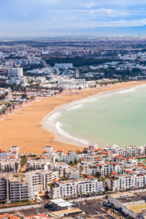 Luftaufnahme einer großen, modernen Küstenstadt, im Bildvordergrund ragen zwei Häuserzeilen mit mehrstöckigen Gebäuden bis ins Meer. Ein goldgelber Sandstrand zieht sich bogenförmig entlang der Stadt, links davon ist die Stadt zu sehen, rechts des Strandes erstreckt sich hellblaues Meer.