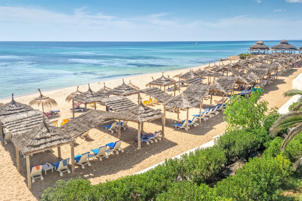 Sandstrand mit Sonnenschirmen aus Bast und blau-weißen Liegen, die in Reihen aufgestellt sind. Im Hintergrund erstreckt sich das türkisfarbene Meer.