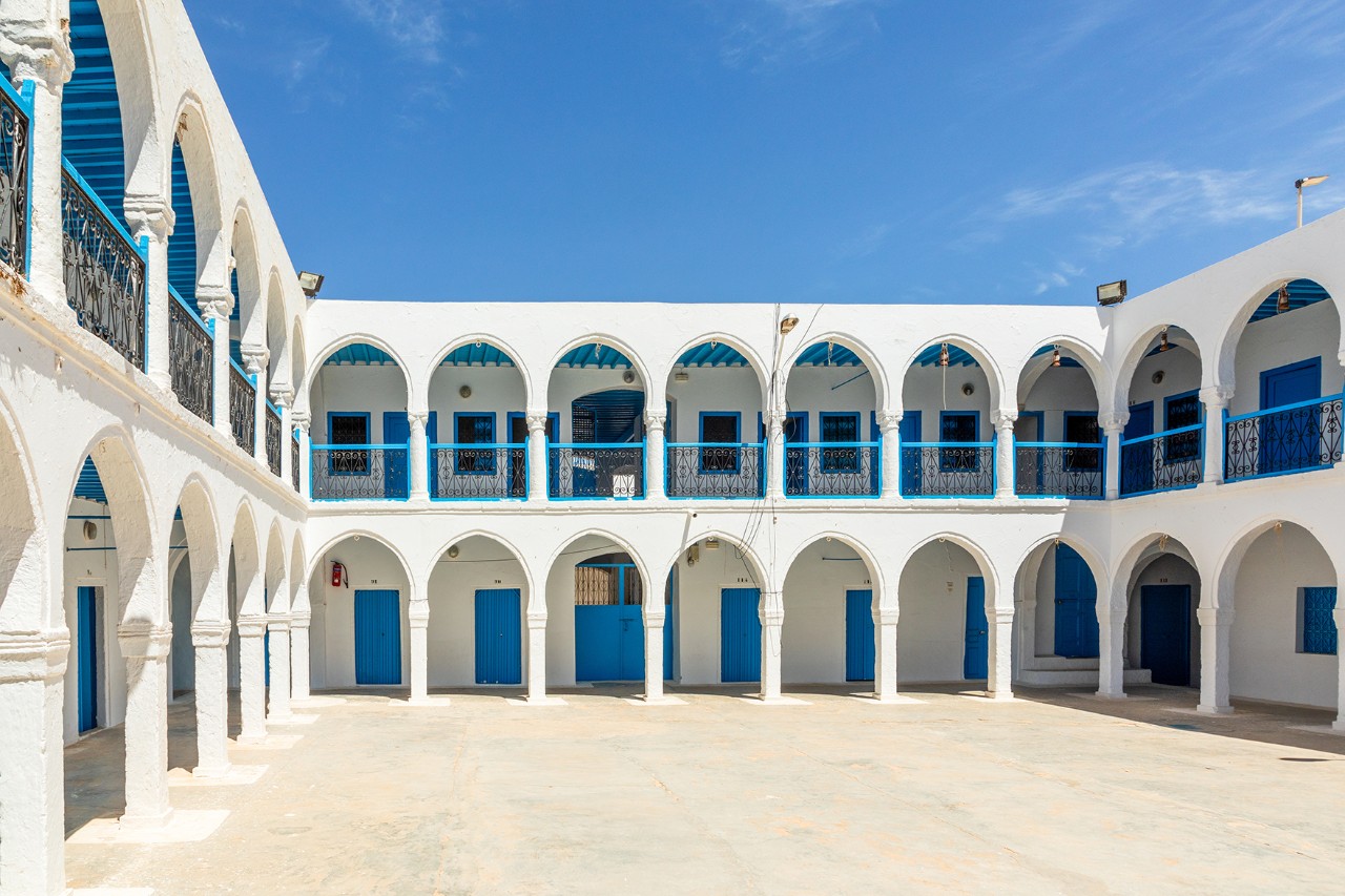 Zweistöckiges weißes Gebäude mit blauen Türen und Fensterläden, Rundbögen und Arkadengängen rund um einen offenen Innenhof.