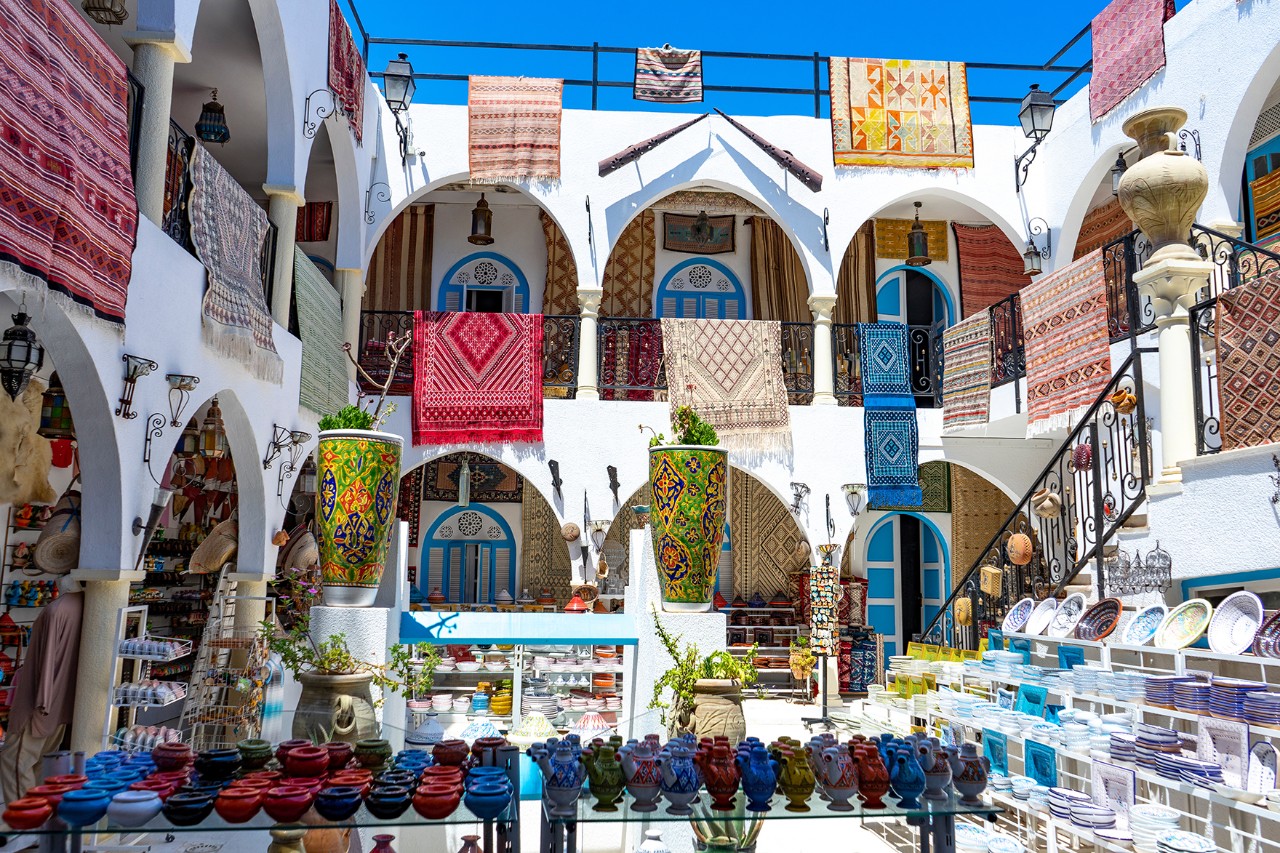 Innenhof eines Souks mit weißen Wänden, Arkaden und blauen Türen. Auf der oberen Etage hängen bunte Teppiche über den Geländern, im Erdgeschoss sind bemalte Keramiken ausgestellt.