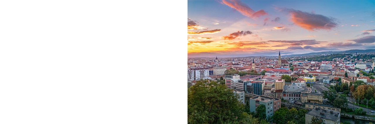 Das Bild zeigt eine größere Stadt im Licht der untergehenden Sonne, mehrere Kirchtürme ragen zwischen anderen Gebäuden hervor und im Hintergrund sind grüne Hügel erkennbar.