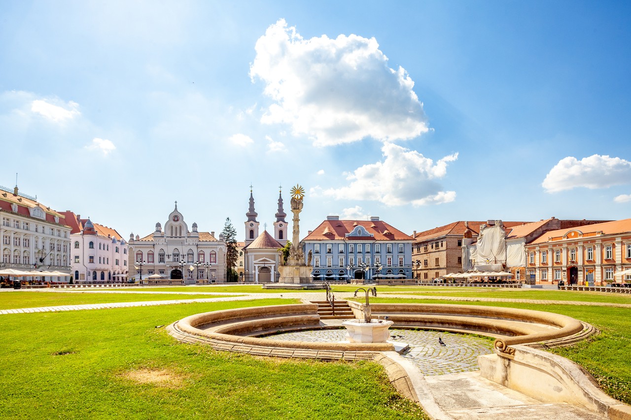 Historischer Platz mit Brunnen, grüner Rasenfläche, barocken Gebäuden und einer Säule unter blauem Himmel mit wenigen Wolken.