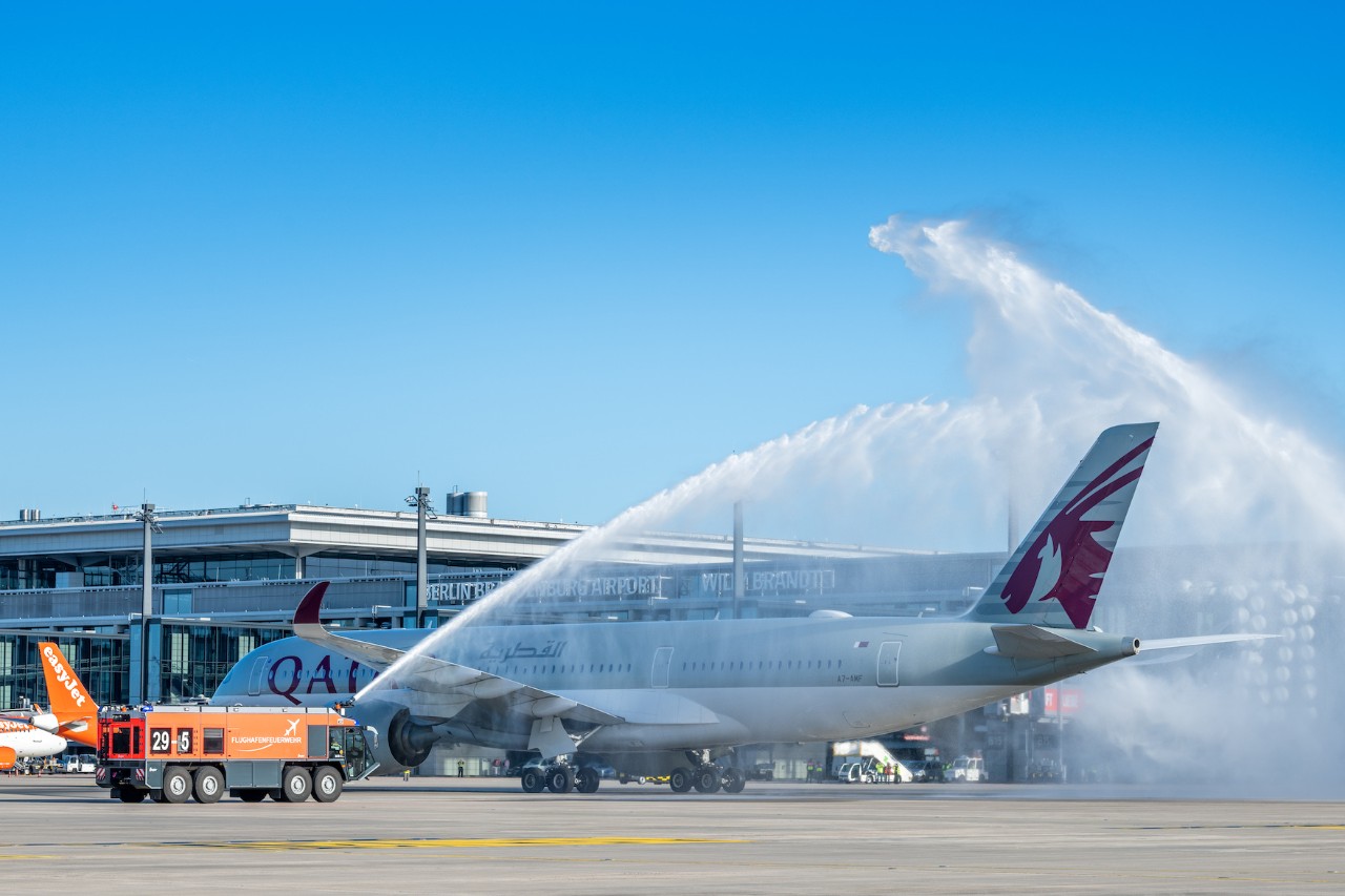 FLugzeug von Qatar Airways wird mit einer Fontäne von 2 Feuerwehrfahrzeugen begrüßt. Dahinter das Terminal 1.