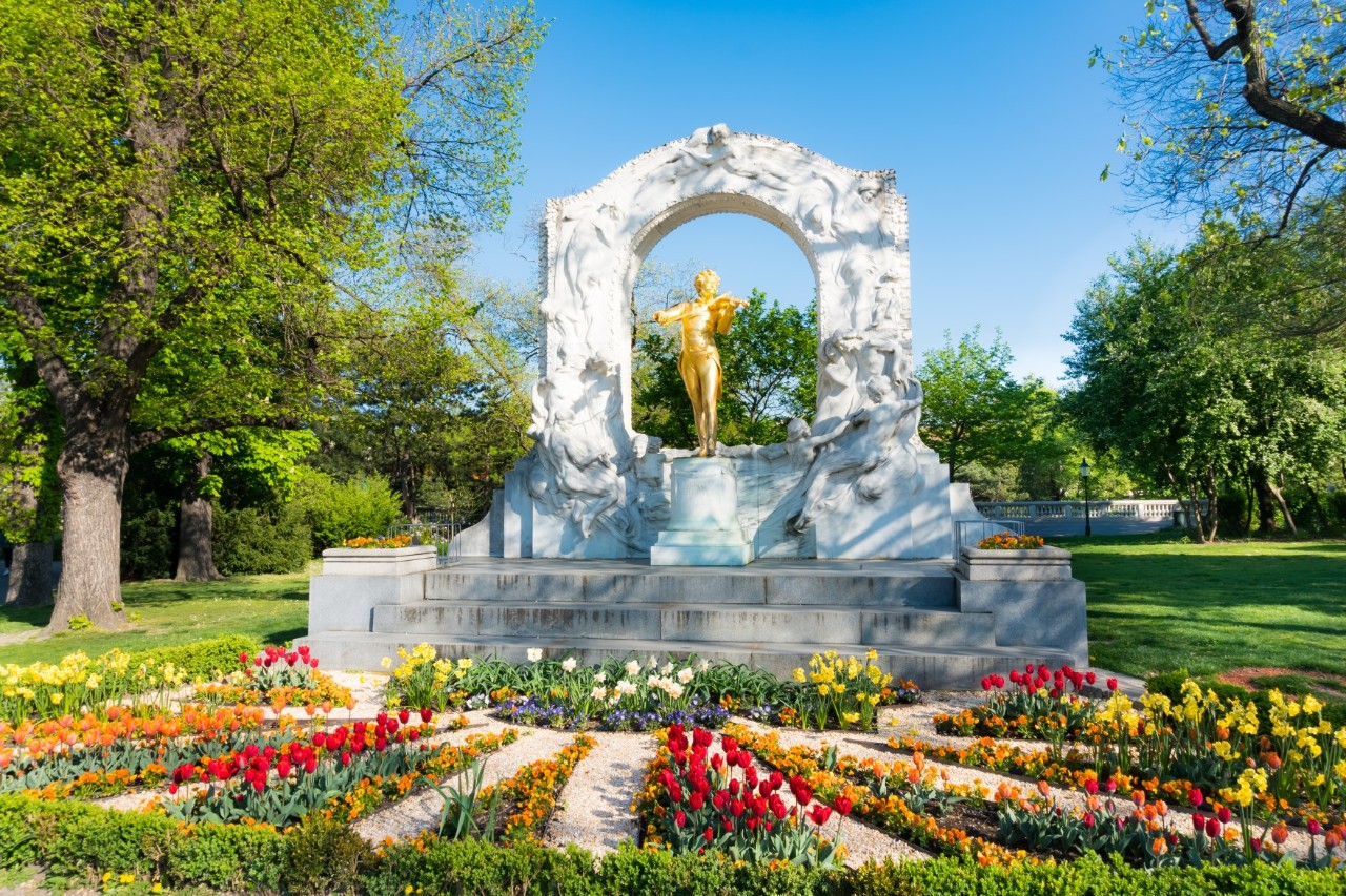 Goldene Strauss-Statue im Stadtpark mit Blumenbeet.