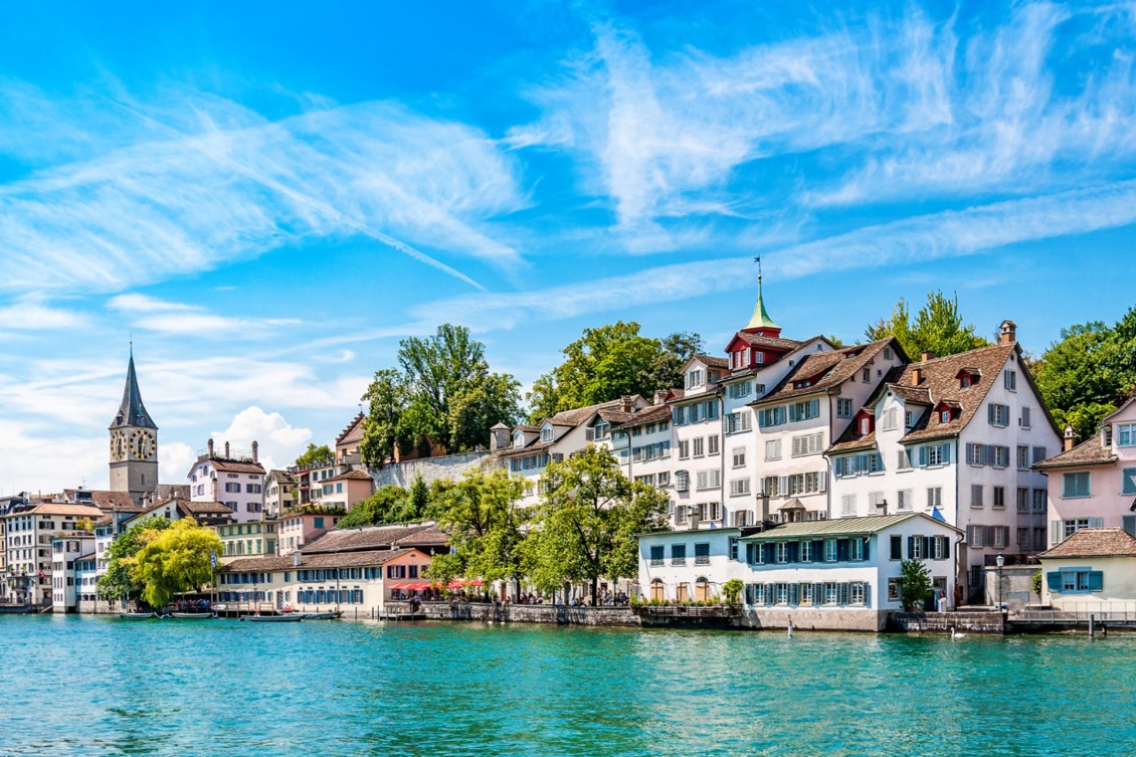 Ein mit eleganten, weißen Häusern bebautes Ufer an einem türkisblauen Gewässer. Zwischen und hinter den Häusern sind grüne Bäume zu sehen. Links im Bild ragt ein Kirchturm über den Häusern empor. Der Himmel ist blau mit wenigen Wolken.
