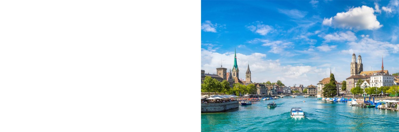 Grünblauer Fluss, der von einem See ausgehend mitten durch eine Stadt verläuft. Auf dem Fluss fahren einige kleinere Boote. An beiden Flussufern sind Bäume, Häuser und Kirchtürme zu sehen. An der vorderen linken Uferseite befindet sich ein belebter Biergarten mit Sonnenschirmen. 
