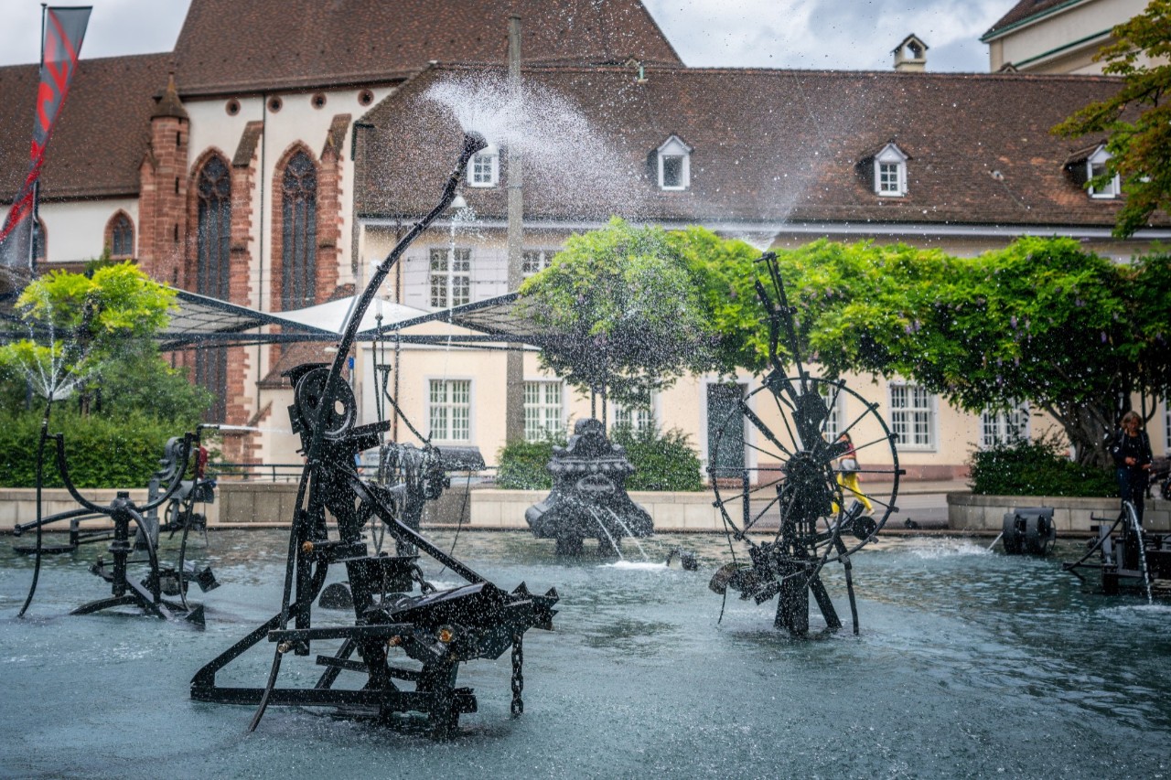 Wassersprühende Kunstfiguren aus Eisen im Wasserbecken vor einem Gebäude und Bäumen 