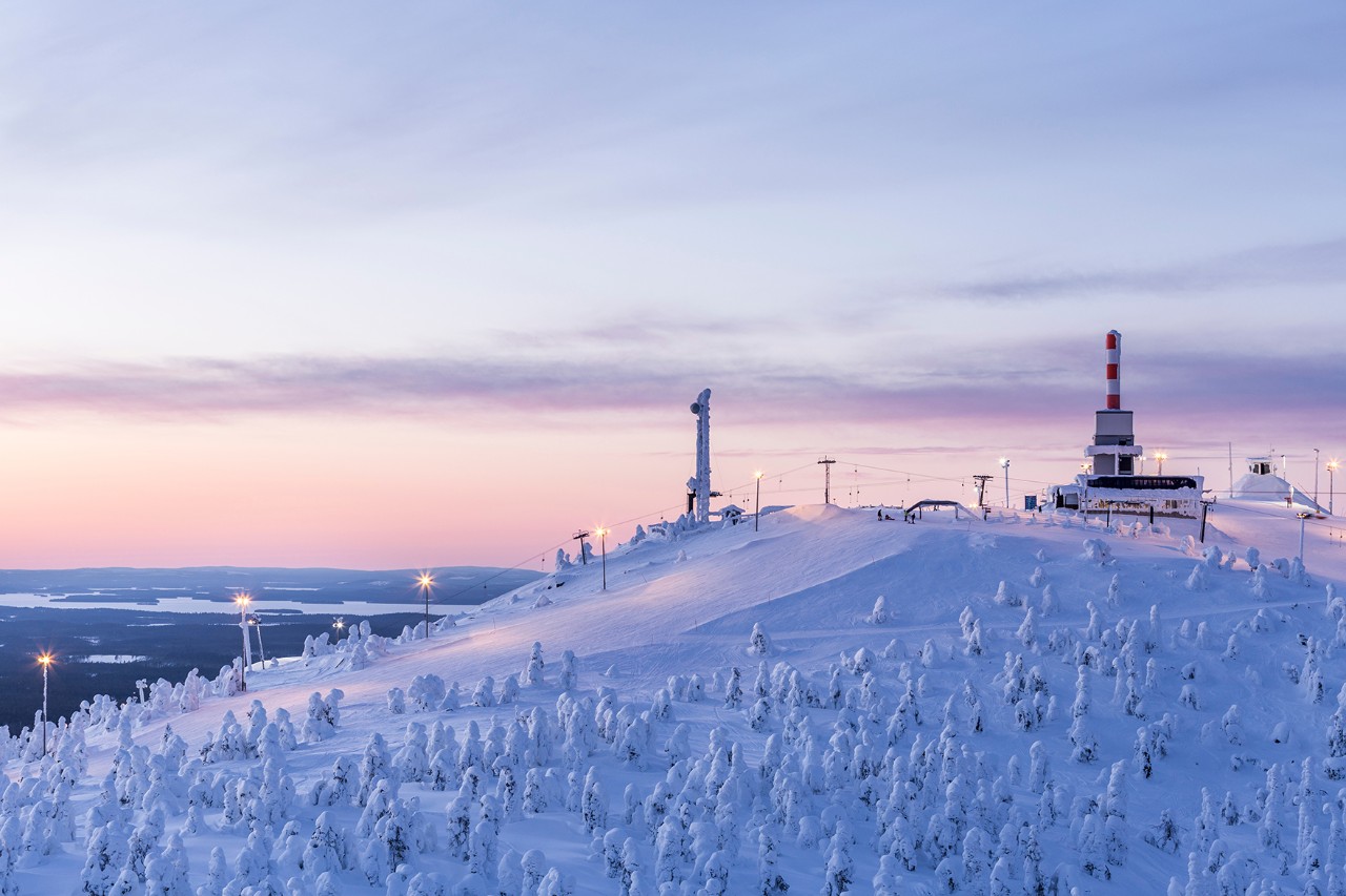 Eine winterliche Schneelandschaft mit kleinen Bäumen und einem Skilift, der entlang einer menschenleeren Skipiste zu einer Bergstation führt. Der Himmel ist teils leicht orange eingefärbt.