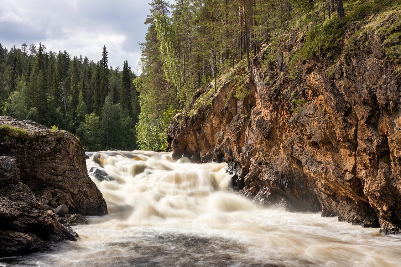 Reißender Fluss mit Wasserfällen zwischen steilen Felsen, umgeben von dichtem Wald.