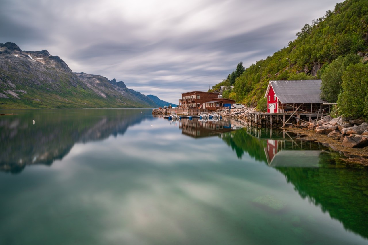 Spiegelglatter Fjord mit roten Holzhäusern und Bergen bei bewölktem Himmel.