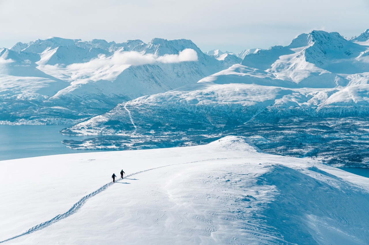 Zwei Skitourengeher auf verschneitem Grat mit Blick auf Fjord und Berge.