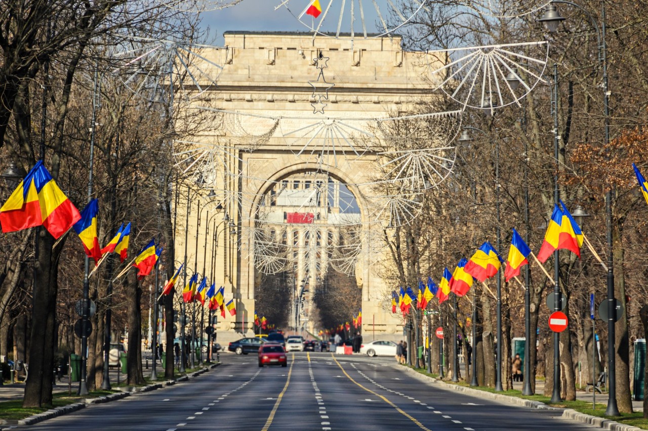 Auf einen Triumphbogen zulaufende vierspurige Allee mit wenigen Autos, links und rechts hohe, gerade, wenig belaubte Bäume, dazwischen Laternenpfähle mit herausragenden rumänischen Fahnen als Schmuck. 