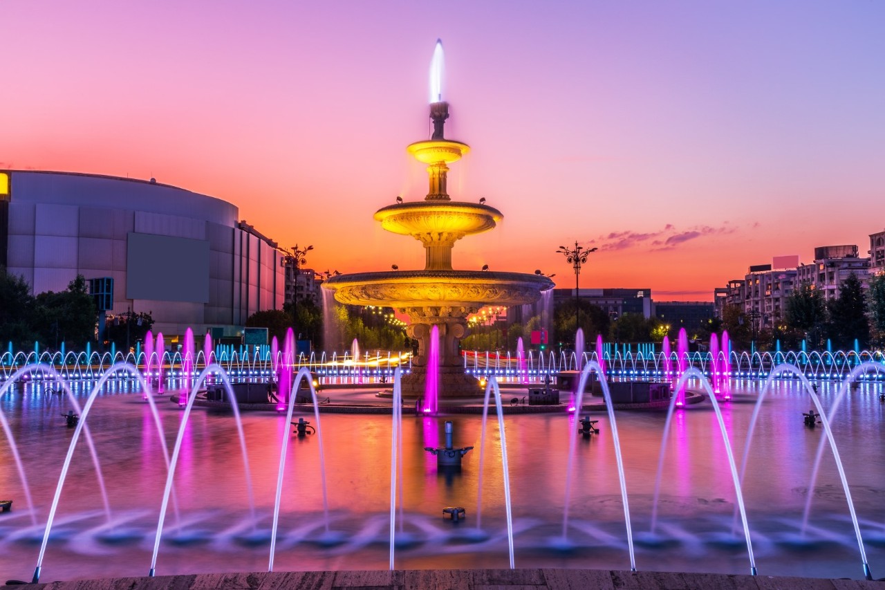 Abendstimmung am lila angestrahlten Springbrunnen mit Häusern im Hintergrund. 