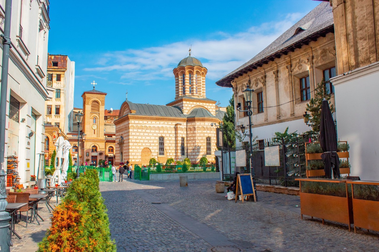 Unbelebter Platz in Bukarest mit Kopfsteinpflaster, zwei Kirchen im Hintergrund, Häusern, Grünpflanzen und Straßencafés.  