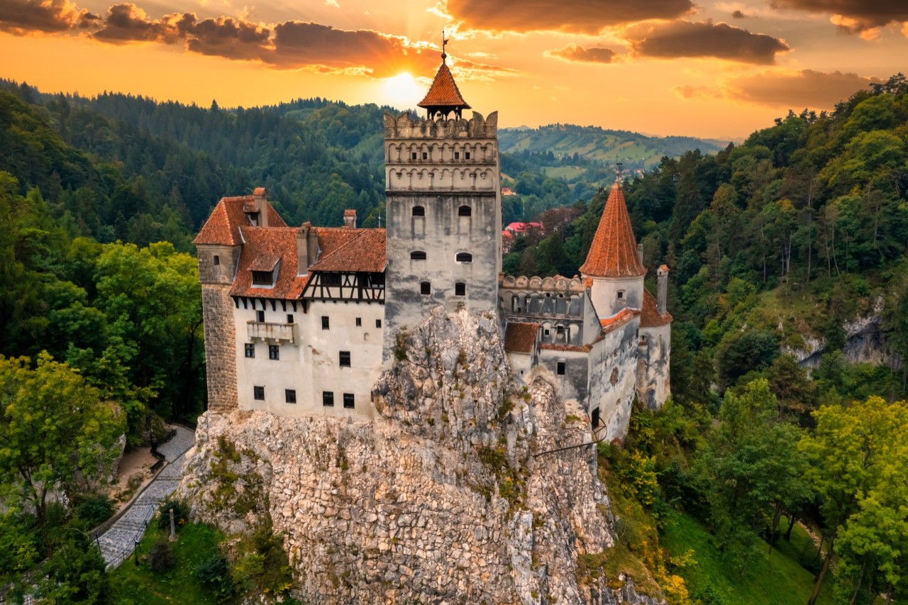 Mittelalterliches Schloss mit Türmen auf einem Felsen, umgeben von hügeligen Wäldern zum Sonnenuntergang. 