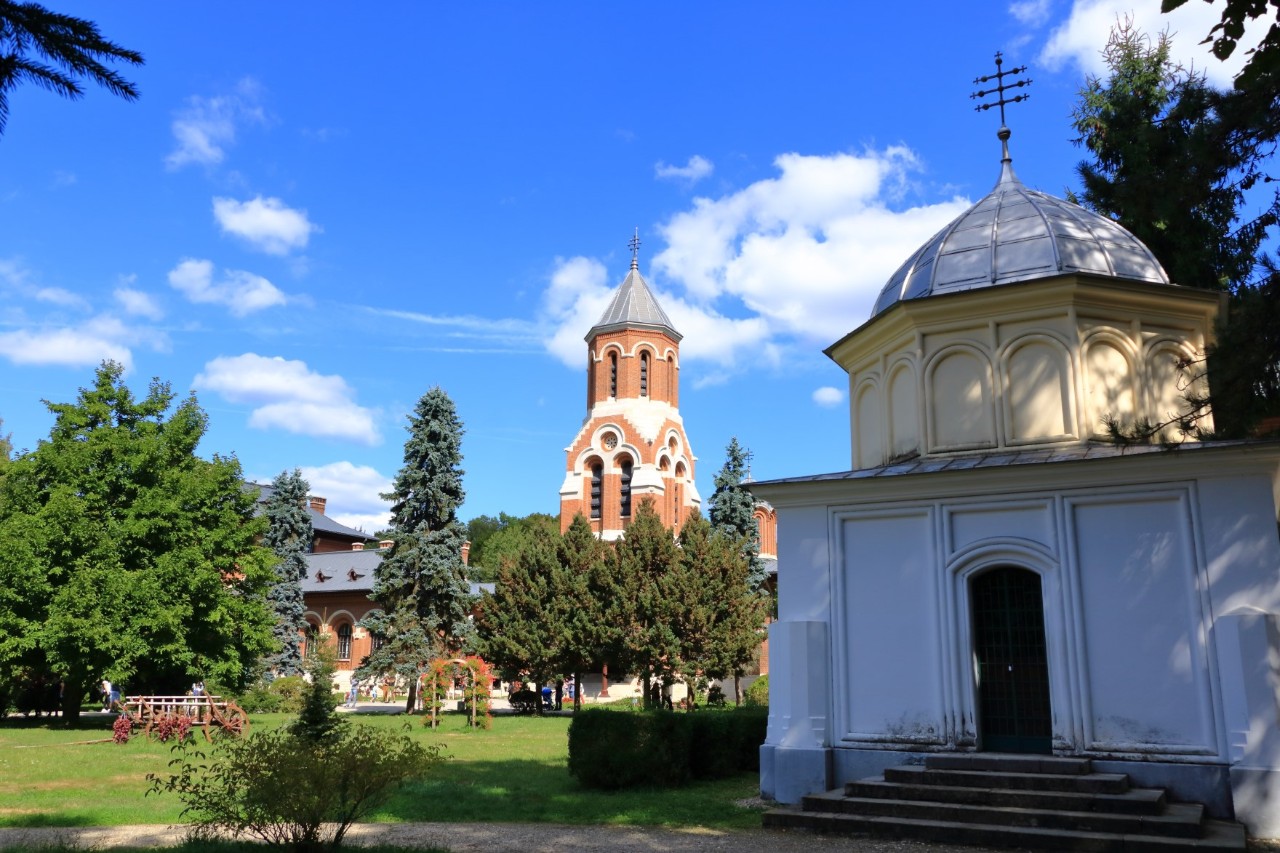 Weiße Kirche im Vordergrund, dahinter Grünanlage mit Bäumen und eine größere, rötliche Kirche.  