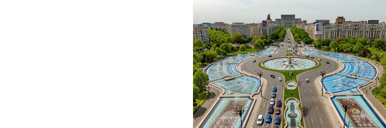 Bukarester Unirii-Platz mit breitem Boulevard, Springbrunnenanlage, Autos, Bäumen, Häusern und dem Parlamentspalast im Hintergrund. 