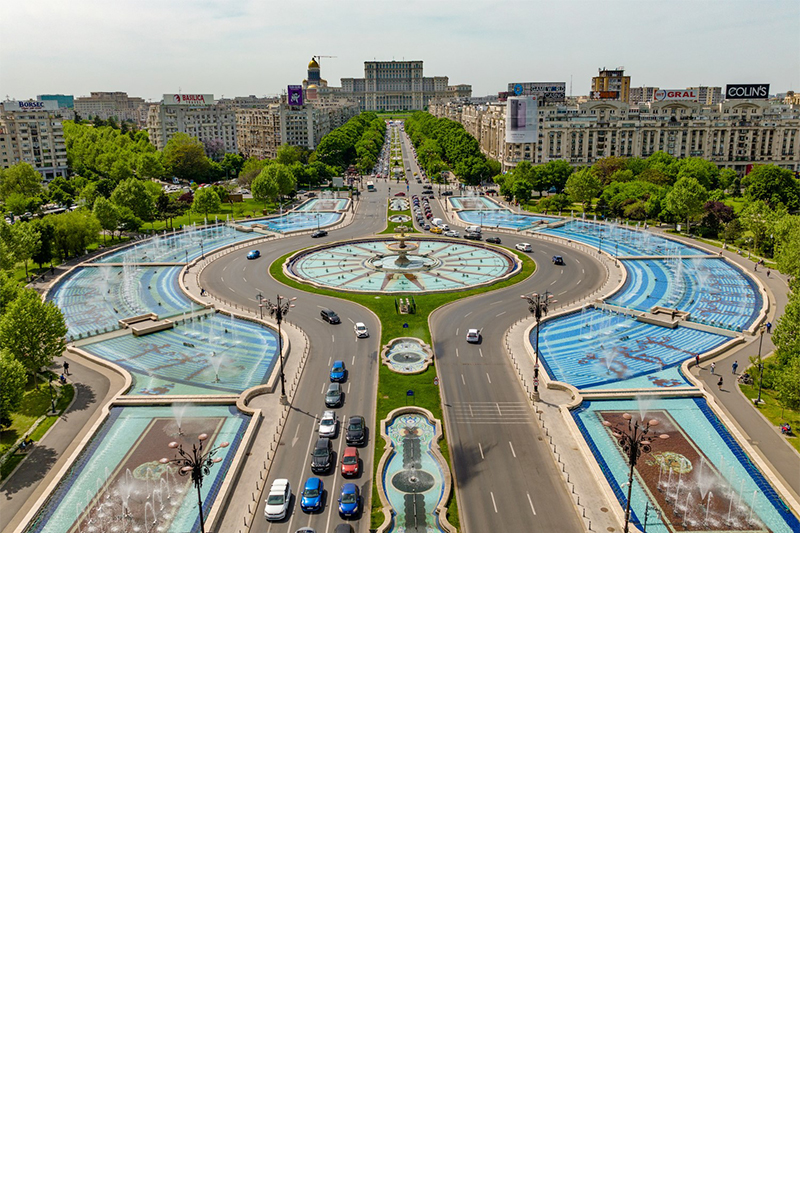 Bukarester Unirii-Platz mit breitem Boulevard, Springbrunnenanlage, Autos, Bäumen, Häusern und dem Parlamentspalast im Hintergrund. 