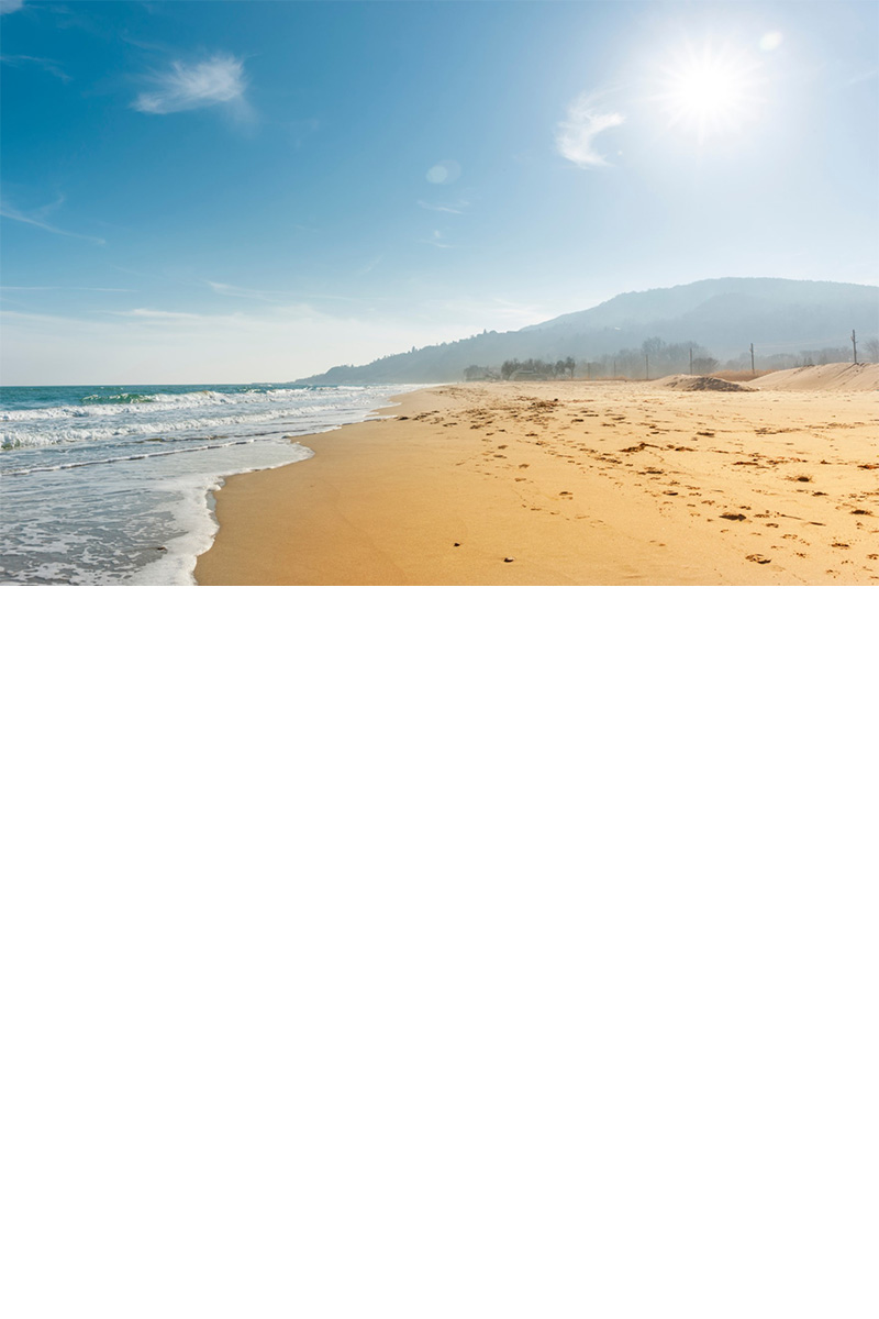 Sandstrand mit ruhigem Meer, dahinter ein sonniger Berg.