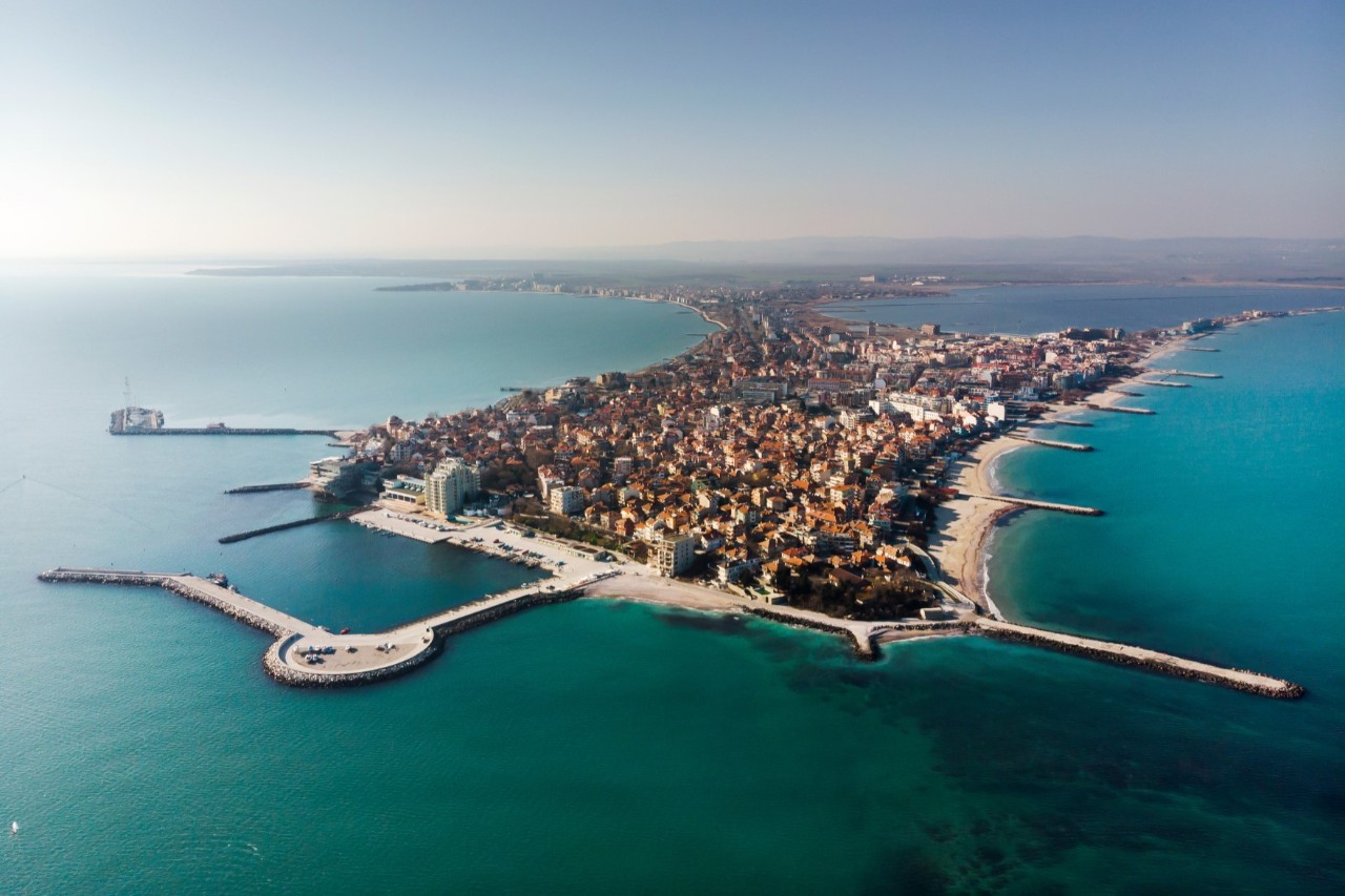 Luftaufnahme einer Halbinsel, die vom Meer umgeben ist. Die Stadt erstreckt sich entlang der Küste mit einer dichten Bebauung aus Wohnhäusern. Strände und Hafenanlagen säumen die Küste.