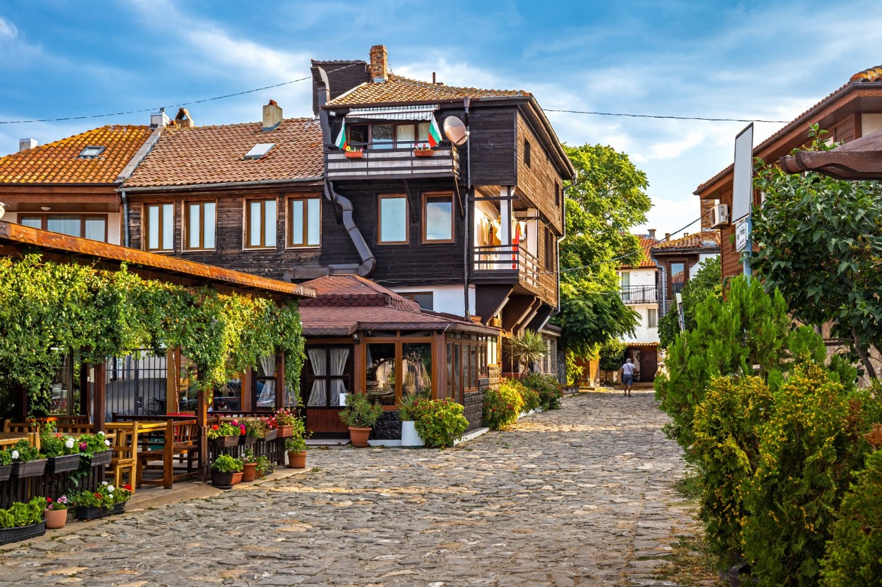 Gepflasterte Straße in einem historischen Stadtteil mit traditionellen Häusern aus Holz und Ziegeln. Im Vordergrund ist ein Restaurant zu sehen, das mit Pflanzen dekoriert ist.