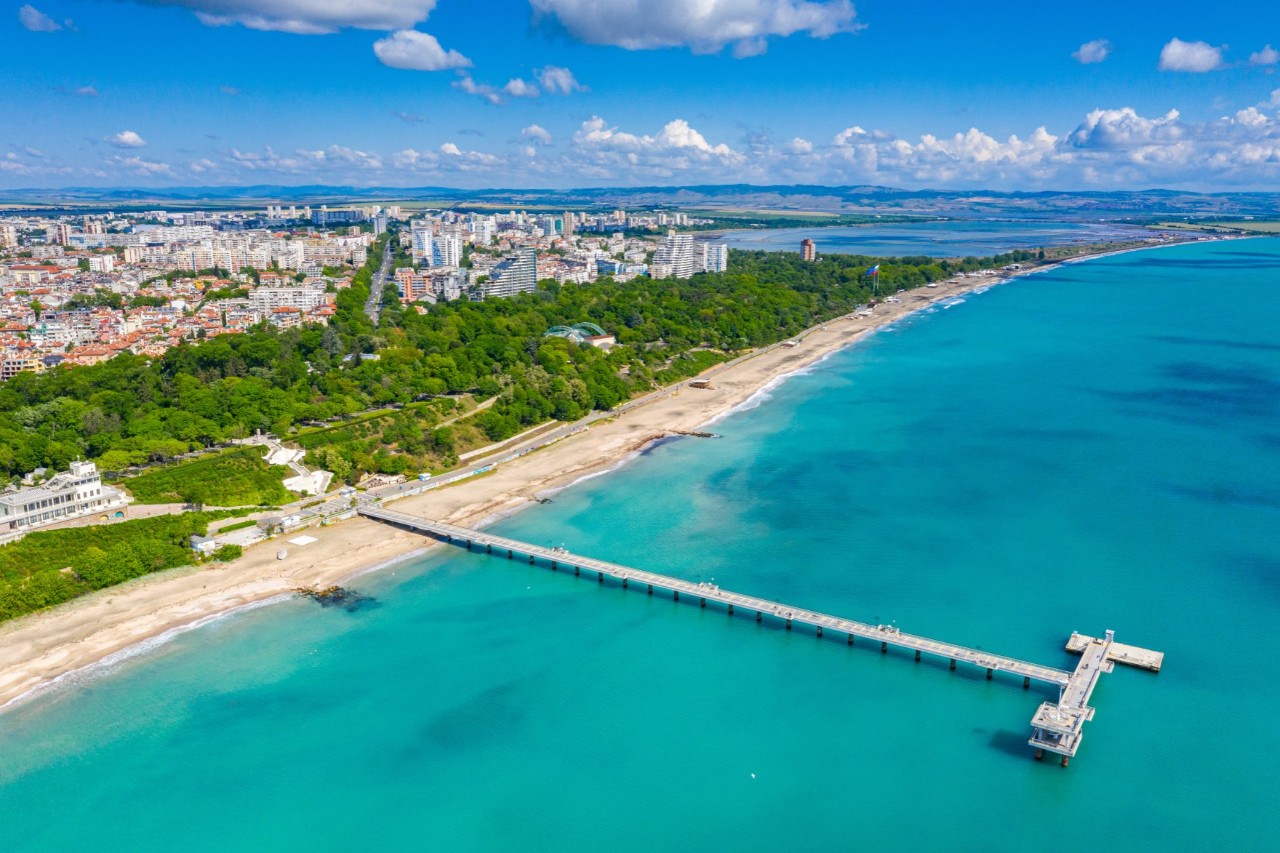 Küstenstadt mit einem langen Pier, der ins türkisfarbene Meer hinausragt. Am Ufer erstreckt sich ein Sandstrand, dahinter eine grüne Parkanlage. Im Hintergrund ist die städtische Bebauung zu sehen.
