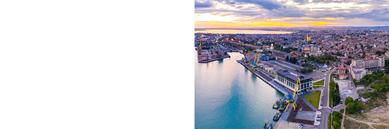 Panoramaaufnahme von Burgas. Links ist der Hafen zu sehen, während rechts ein Sandstrand und das Schwarze Meer erkennbar sind. In der Bildmitte erstreckt sich die Stadt mit Gebäuden und Grünflächen. 