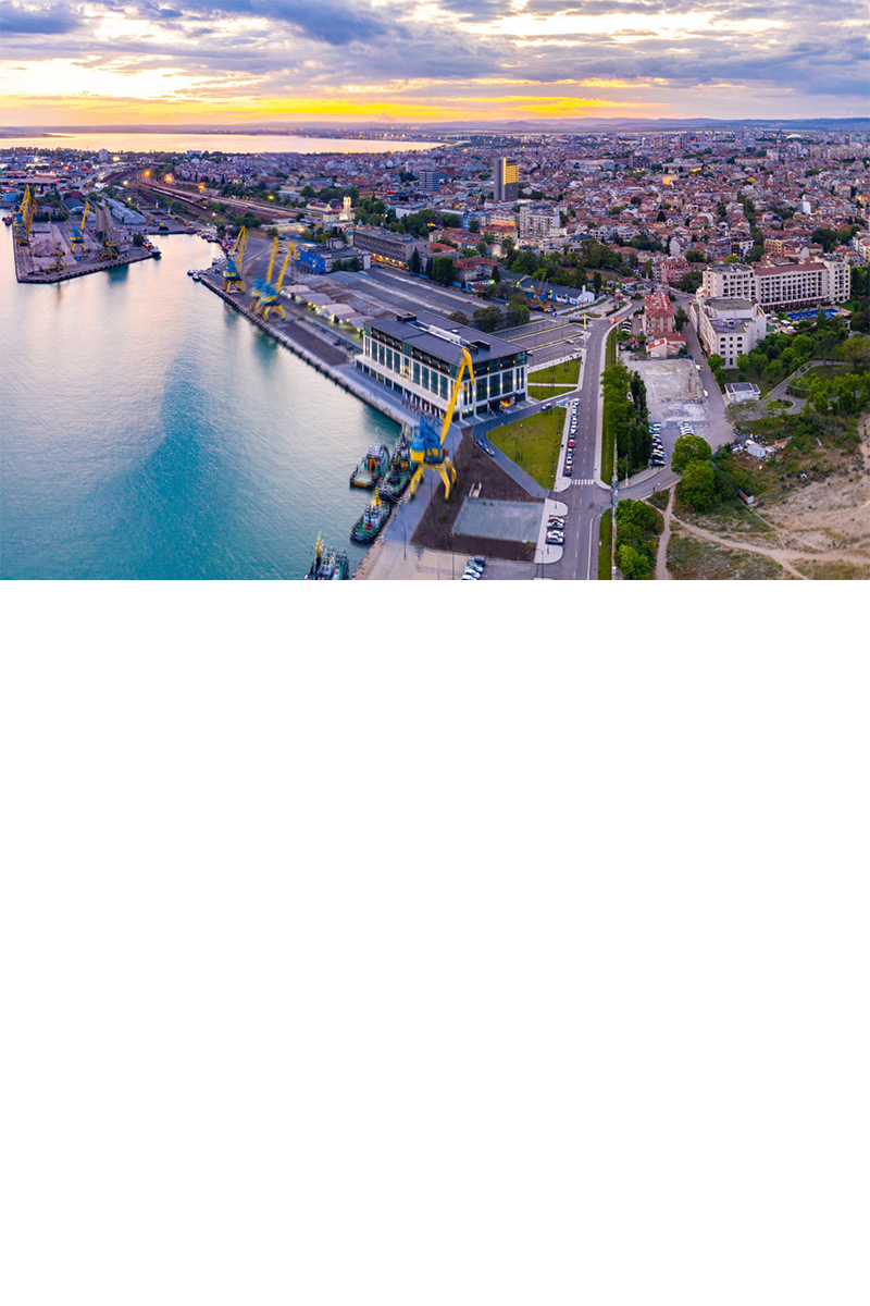 Panoramaaufnahme von Burgas. Links ist der Hafen zu sehen, während rechts ein Sandstrand und das Schwarze Meer erkennbar sind. In der Bildmitte erstreckt sich die Stadt mit Gebäuden und Grünflächen.