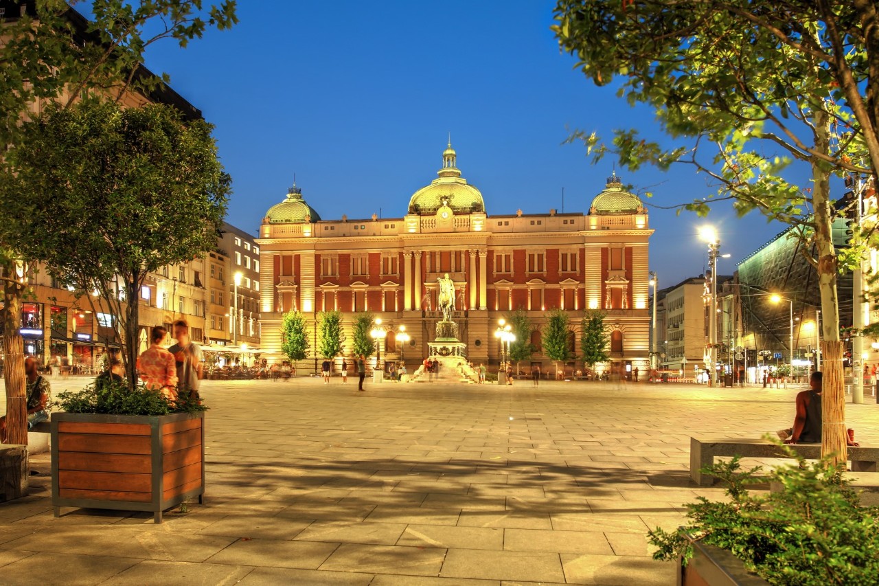 Ein Platz bei abendlicher Beleuchtung. Im Bildvordergrund stehen zu beiden Seiten in Kübel gepflanzte Bäume. Im Bildzentrum ist ein großes historisches Gebäude mit roter Fassade und drei Türmen zu sehen. Vor dem beleuchteten Gebäude stehen eine Statue, rechts und links daneben sind weitere Bäume.