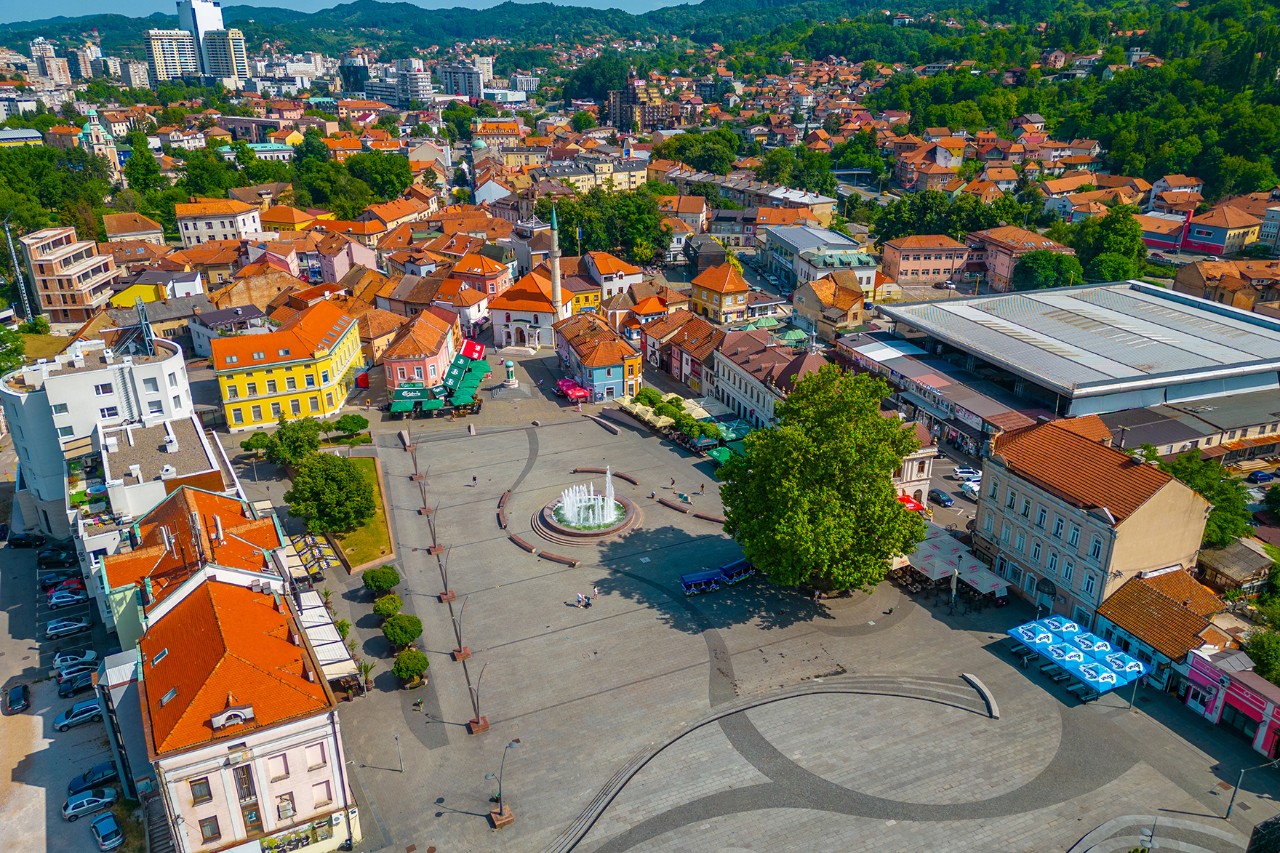 Blick von oben auf einen wenig belebten Stadtplatz mit Brunnen in der Mitte, umgeben von bunten Häusern mit roten Dächern. Im Hintergrund sind Hügel, Wohngebiete sowie einige größere Gebäude und viel Grün.