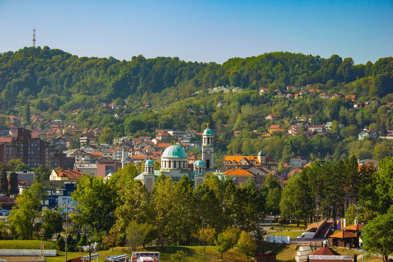 Ortschaft in hügeliger, grüner Landschaft mit Häusern und einer Kirche mit grünen Kuppeln im Zentrum.