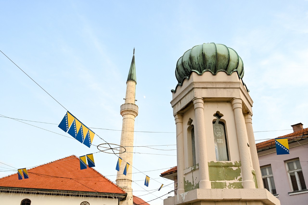 Detail einer Stadtansicht mit dem Minarett einer Moschee und einem Turm mit grüner Kuppel; zwischen den Häusern mit roten Dächern hängen kleine bosnische Flaggen über der Straße.