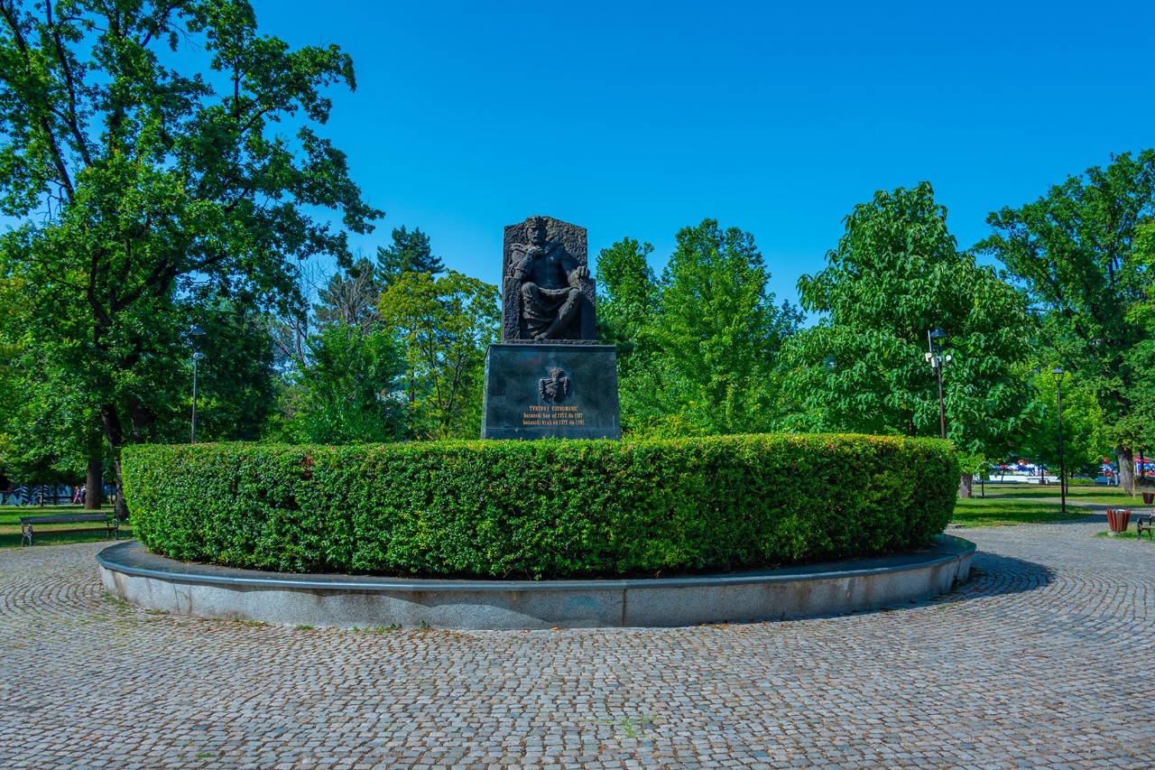 Schwarzes Denkmal mit sitzender Statue auf einem Sockel, umgeben von einer runden grünen Hecke in einem Park mit Bäumen und gepflastertem Weg.