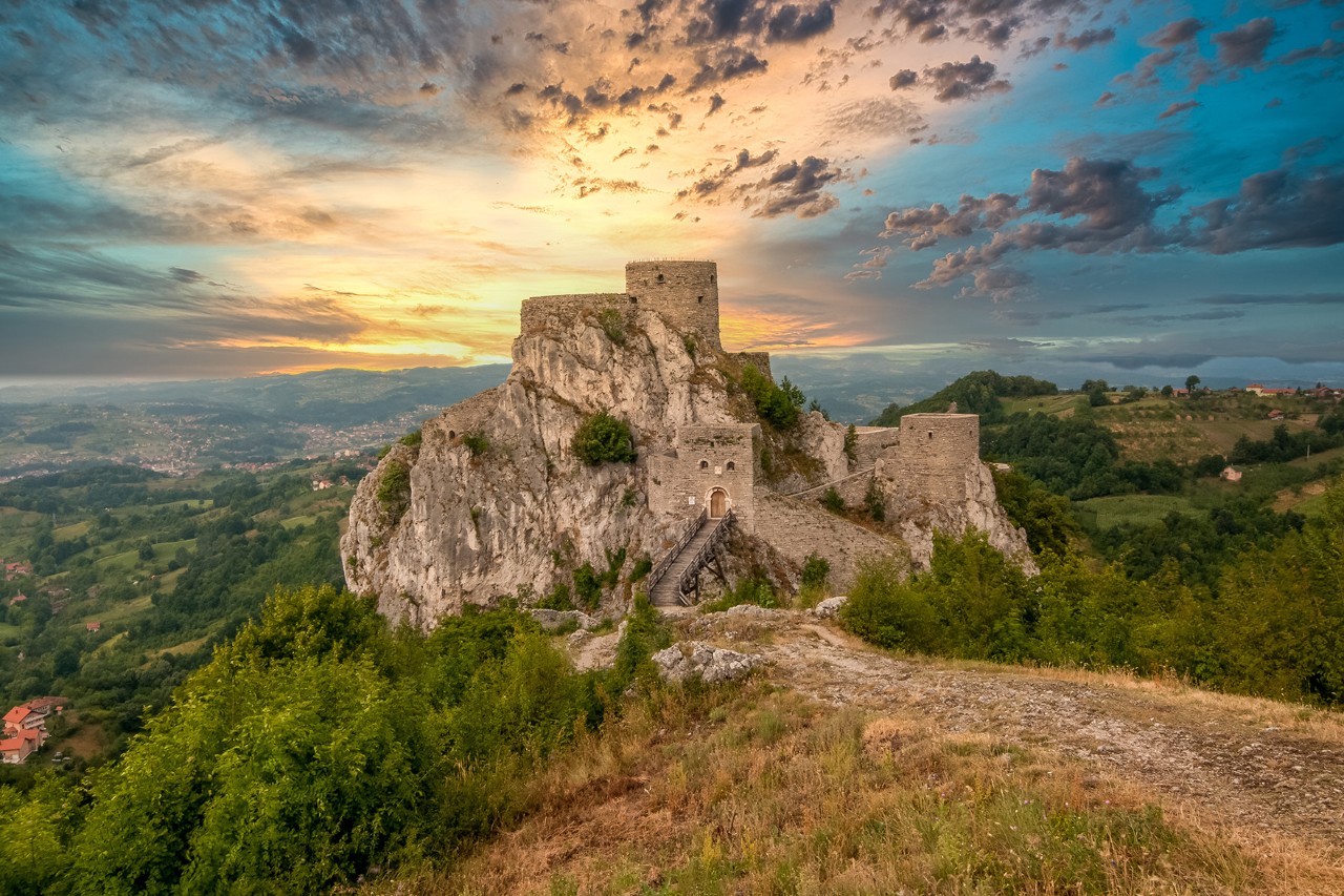 Mittelalterliche Burg auf einem felsigen Hügel, umgeben von grüner Landschaft; dramatischer Sonnenuntergang mit farbigen Wolken am Himmel.